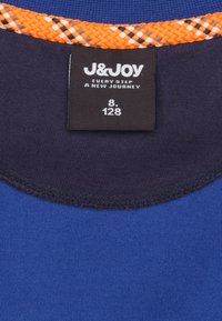 Vêtement en textile bleu avec un col côtelé marine et un accent tissé orange au niveau de l'encolure ; l'étiquette comporte le nom de la marque et les informations de taille.