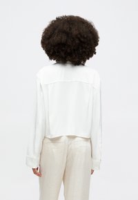 White, cropped long-sleeve shirt with a smooth texture, featuring a back yoke detail. Paired with light beige trousers with back pockets.