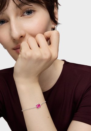 Młoda osoba z krótkimi brązowymi włosami i srebrnymi kolczykami w kształcie koła, opierająca podbródek na dłoni, ubrana w ciemnoczerwoną koszulę i srebrną bransoletkę z różowym zawieszką.