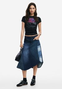 Falda de mezclilla oscura con dobladillo asimétrico, bolsillo de parche en contraste y frente con cinco botones. Combinada con una camiseta gráfica negra y zapatillas.