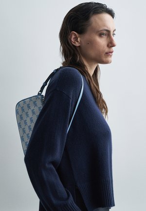 Canvas backpack in light blue featuring a pattern of dark blue logo lettering, worn on the back. Model in a navy sweater, side profile view.