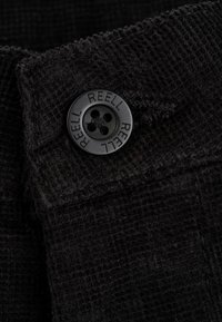 Black corduroy fabric showcasing a textured surface. Features a black button with four holes and "REELL" engraved around the edge.