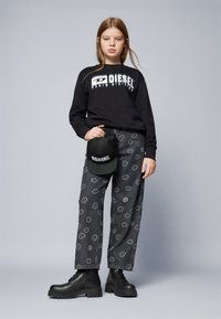 Felpa nera con logo "Diesel", pantaloni grigi con motivo a faccina sorridente, cappellino nero e stivali neri robusti, tutti su uno sfondo semplice.