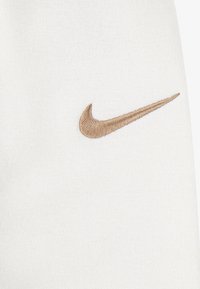 Balts audums ar teksturētiem adījuma rakstiem, kurā sānos ir zelta izšūta Nike swoosh emblēma. Vienkāršs un minimālistisks dizains.