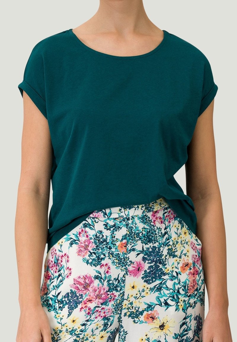 Personne portant un haut à manches courtes bleu sarcelle foncé et un pantalon blanc avec un imprimé floral multicolore, debout sur un fond uni.