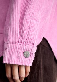 Ružová košela z kordu so svislým ribbovaným vzorom, so strieborným kruhovým logom na gombíku a prešitými manžetami. Na pozadí je viditeľná hnedá kordová textúra.