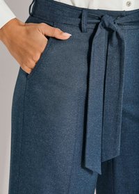Hand in pocket van blauwe hoge taille broek met stoffen riem, die in een strik is geknoopt, gecombineerd met een wit shirt met een decoratieve knop.