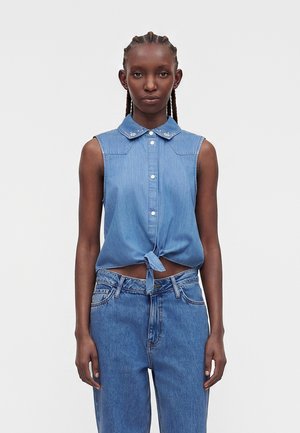 Vrouw met gevlochten haar die een mouwloos gebonden blauwe denim blouse met geborduurde kraag en hoge taille blauwe spijkerbroek draagt, staand tegen een witte achtergrond.