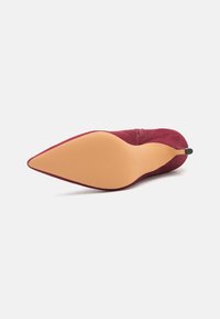 Zapato de tacón alto de ante rojo con punta afilada y suela beige lisa. Presenta una textura sutil y un pequeño acento negro en el tacón.