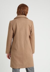 Lång, kamelfärgad kappa i mjukt tyg. Har en snibbkrage och rak skärning som når mitt på låret. Diskret texturerad yta.