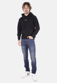 Felpa nera in cotone con cordini e tasca frontale, abbinata a jeans in denim blu e sneakers bianche con logo swoosh nero.