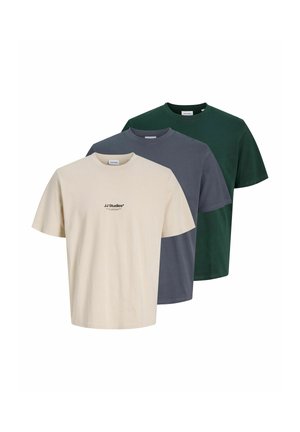 Jeune homme debout portant un t-shirt beige à manches courtes, un pantalon noir et des baskets noires et blanches sur un fond clair uni.