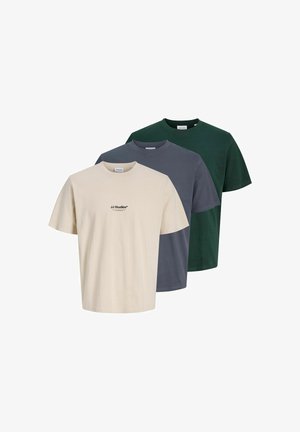 Jeune homme debout portant un t-shirt beige à manches courtes, un pantalon noir et des baskets noires et blanches sur un fond clair uni.