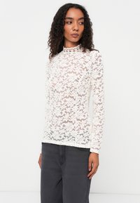 Fehér csipkés, hosszú ujjú felső virágmintával és csipkés gallérral; áttetsző anyag; sötét szürke nadrág fölé viselve.