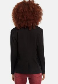 Mujer con cabello rizado castaño rojizo que lleva un blazer negro ajustado y jeans rojos, vista desde atrás contra un fondo blanco liso.