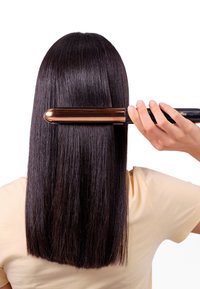Lisseur avec une poignée noire élégante et une plaque rose dorée brillante, glissant à travers de longs cheveux noirs lisses sur fond blanc.