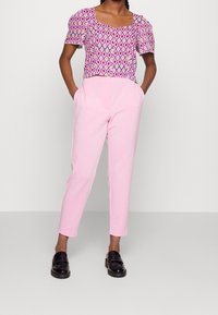 Top corto a fantasia nei colori rosa, bianco e viola con forme a diamante, abbinato a pantaloni a sigaretta rosa chiaro. Le scarpe nere senza lacci completano il look.
