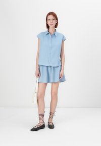 Camicia senza maniche in denim azzurro chiaro con bottoni davanti, abbinata a una gonna coordinata. Scarpe nere basse con fasce incrociate e borsa bianca.