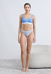 Ανοιχτογαλάζιο ασύρματο σουτιέν με λευκή ζώνη με λογότυπο, συνδυασμένο με γκρι σλιπ με πουά σχέδια ψηλής κοπής. Λείο ύφασμα, εφαρμοστό σχέδιο και ελάχιστες λεπτομέρειες.