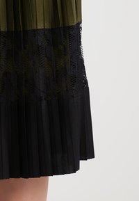 Gonna midi plissettata con una parte superiore verde oliva e una parte inferiore in pizzo nero testurizzato, caratterizzata da pieghe verticali e dettagli intricati.