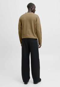 Persona de piel oscura vistiendo un suéter marrón, pantalones de pierna ancha negros y zapatos negros, de pie de espaldas contra un fondo claro liso.