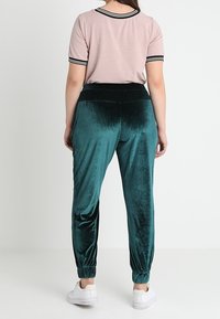 Jogging en velours teal avec des poignets élastiques, associé à un haut ajusté rose avec des accents rayés noir et blanc au niveau du col et des manches.