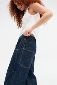 Jeans ampi in denim scuro con vita alta, caratterizzati da cuciture gialle e molteplici tasche, abbinati a un top bianco senza maniche.