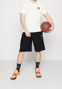 Camiseta blanca con un logo colorido, pantalones cortos negros y zapatillas de baloncesto naranjas. El conjunto se complementa con calcetas negras que llevan un logo.