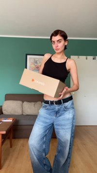 Κουτί Zalando στο χέρι, φορώντας ένα μαύρο αμάνικο μπλουζάκι και φθαρμένα μπλε παντελόνια φάρδους. Το φόντο περιλαμβάνει πράσινους τοίχους και έναν καναπέ.