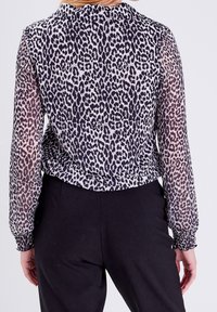 Transparente Bluse mit einem Schwarz-Weiß-Leopardenmuster, gerafften Manschetten und hohem Kragen, kombiniert mit schwarzen Hosen.