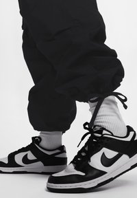 Černobílé tenisky Nike s texturovaným koženým svrškem, šněrováním a akcenty loga, které jsou v kombinaci s volnými černými joggingovými kalhotami a bílými žebrovanými ponožkami.