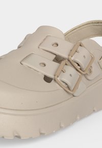 Gros plan d'une chaussure beige à enfiler avec deux sangles réglables et des boucles rectangulaires assorties sur une semelle épaisse texturée.