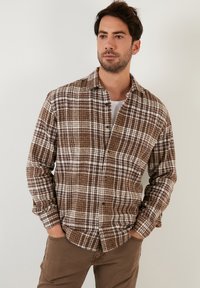 Camicia a quadri marrone e bianca realizzata in tessuto testurizzato con chiusura a bottoni e colletto; il modello la indossa aperta sopra una t-shirt bianca.