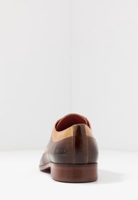 Chaussure en cuir marron avec des accents beige contrastants, bout arrondi et texture lisse. Présente un logo de marque embossé sur le talon.