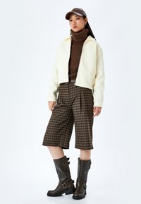 Krämfärgad kort jacka, brun stickad turtleneck, bruna rutiga vida shorts och mörkbruna knähöga stövlar med spännen.