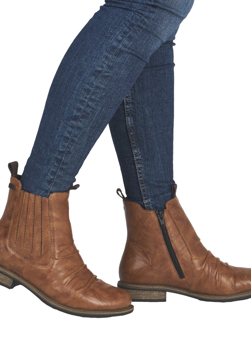 Jambes portant un jean skinny bleu foncé et des bottines en cuir marron avec des fermetures éclair et des détails de couture.