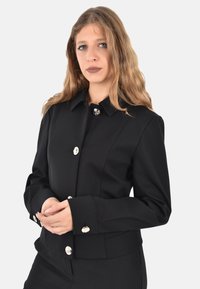 Giacca nera realizzata in tessuto liscio, caratterizzata da un design con colletto, bottoni argentati e una silhouette aderente con maniche sartoriali.