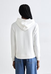 Hvid hoodie med en blød tekstur, der har en front kangaroo-lomme, fastgjort hætte og ribbede manchetter. Båret over blå denimbukser.
