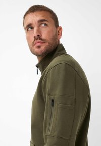 Felpa verde oliva a metà zip realizzata in materiale morbido, con colletto alto, tessuto testurizzato e tasca con zip sulla manica.