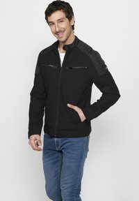 Jachetă neagră cu panouri matlasate pe umeri, buzunare cu fermoar pe piept și buzunare laterale. Fabricată dintr-un material moale și prezintă un guler în picioare.