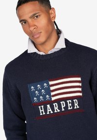 Granatowy sweter robiony na drutach z haftowaną amerykańską flagą i napisem "HARPER". Kołnierz ma kontrastującą koszulę w paski pod spodem.