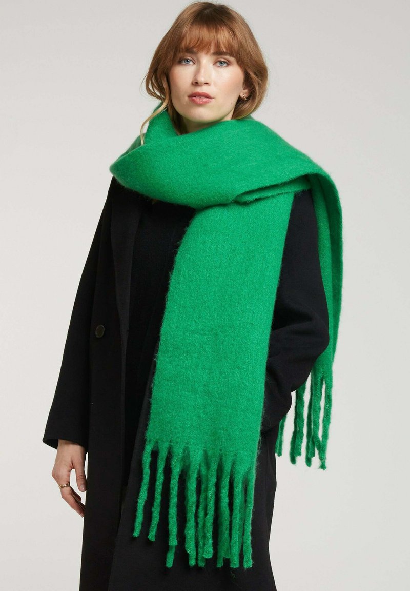 Écharpe en laine verte avec des bords fringés, portée sur un manteau noir. Le tissu texturé est doux et chaud, de couleur vive et unie.