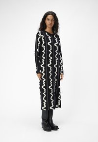 Robe-sweater noire à manches longues, ornée de motifs en zigzag blancs. Confectionnée dans un tissu texturé. Associée à des bottes noires montantes.