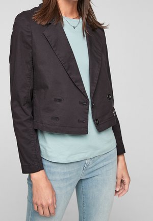 Vrouw draagt een zwart cropped blazer over een lichtblauw shirt en lichtgewassen spijkerbroek, kijkt recht vooruit met haar rechterhand ontspannen langs haar zij.