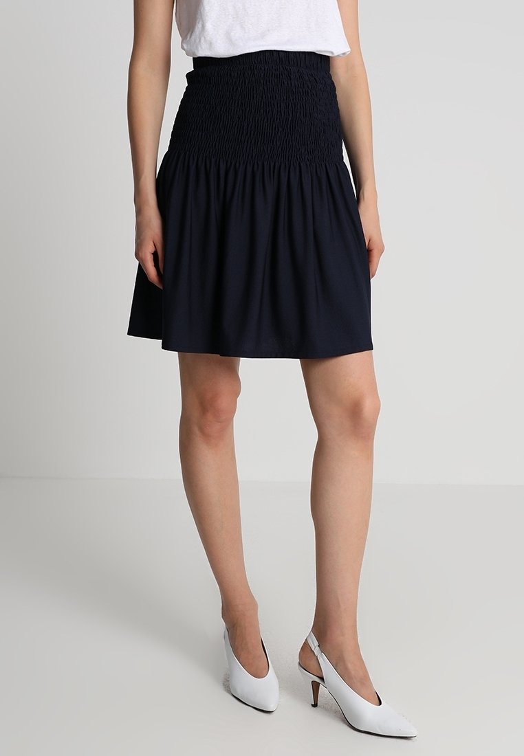 Femme portant une jupe noire froncée, un haut blanc et des talons blancs à bride arrière, debout contre un fond gris uni.