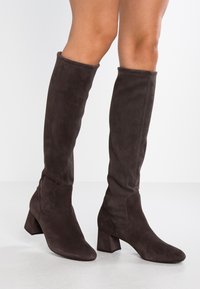 Bottes hautes jusqu'au genou en daim marron, avec un talon carré, un bout arrondi et des coutures ton sur ton. Texture lisse avec une coupe ajustée autour de la jambe.
