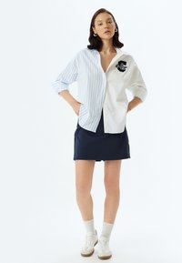 Blau gestreiftes und weißes Hemd mit Knopfleiste und besticktem Patch, kombiniert mit einem marineblauen Rock und weißen Sneakers mit hellgrauen Socken.