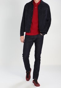 Veste en daim noir superposée à un polo rouge, assortie à un jean foncé et des baskets bourgogne. Design minimaliste avec des poches avant sur la veste.