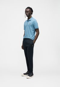 Polo azul com um logótipo amarelo, combinada com calças de fato de treino escuras e sapatilhas pretas de cano alto. Tecido suave com design clássico de colarinho.