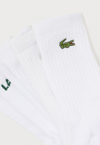 Trys baltų trumpinių kojinių poros su žaliai siuvinėtų krokodilo logotipų juostelėmis, išdėstytos skleidžiasi ant baltos fono.